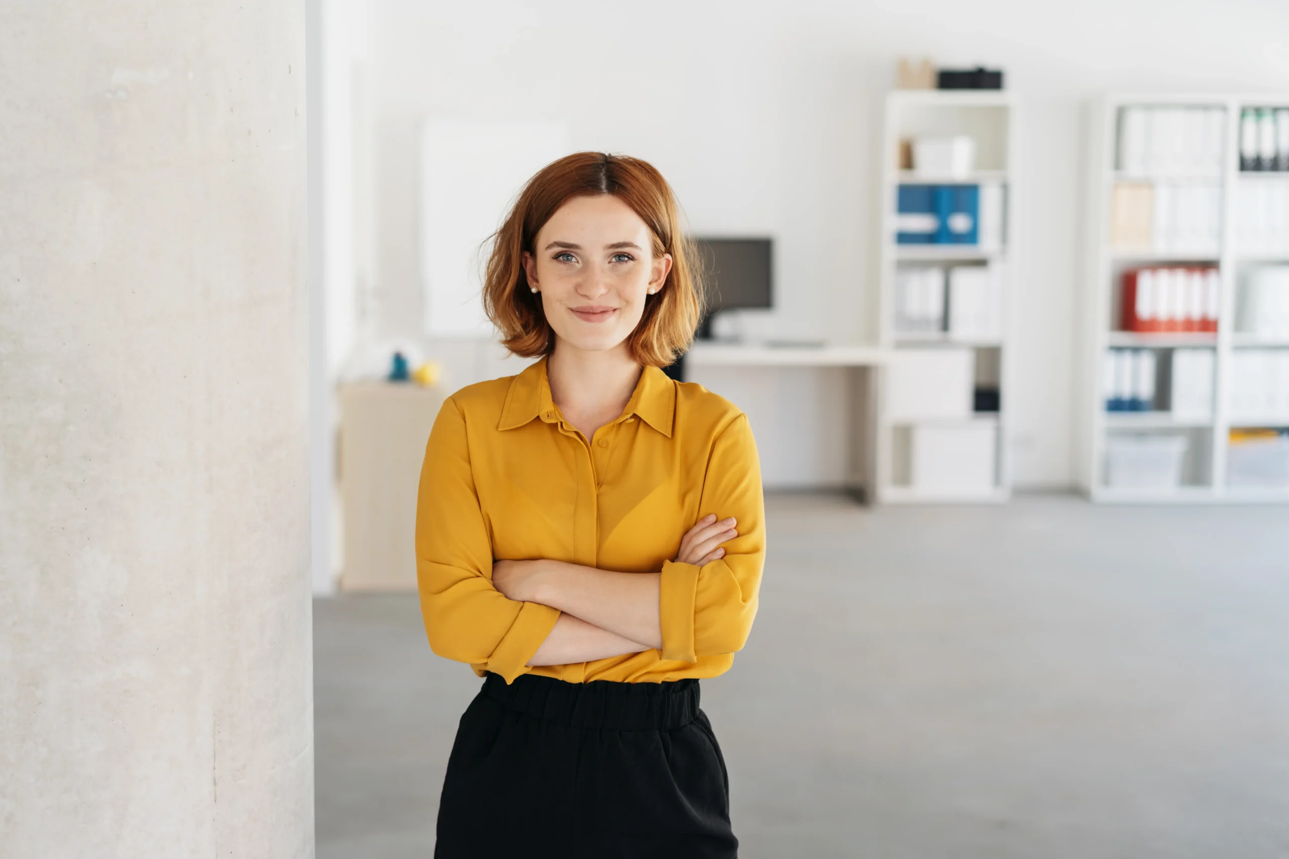 Eine Frau steht in einem Büro, Arme verschränkt, Blick zur Kamera.